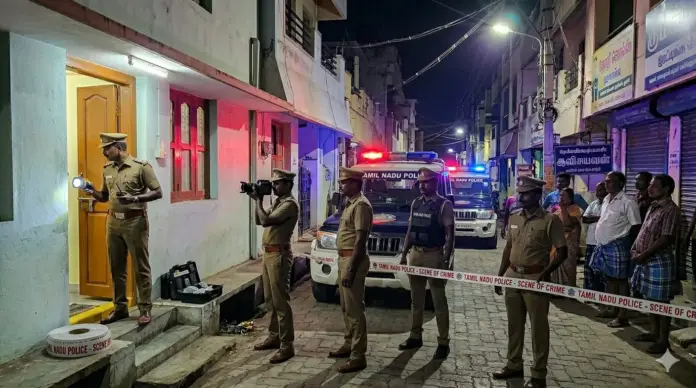 A realistic Tamil news style crime scene illustration showing a police raid at a suspicious rental house in a narrow street in Coimbatore at night. Indian police officers in plain clothes entering a modest house, one police jeep parked outside, tense atmosphere, flashing red and blue lights, local residential area, worried neighbors watching from a distance, no explicit content, no inappropriate visuals, cinematic news coverage style, dramatic lighting, South Indian urban setting, highly realistic, breaking news thumbnail style.